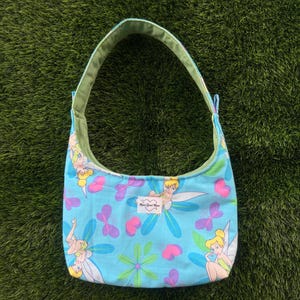 May include: A light blue shoulder bag with a floral and fairy print. The bag features a green strap and lining. The design includes fairies, flowers, and hearts. The text "Main Street Mouse" is visible on the bag.