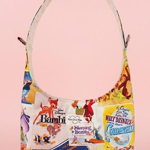 May include: A hobo-style shoulder bag featuring a colourful collage of vintage Walt Disney movie posters including Bambi, Sleeping Beauty, Cinderella, Dumbo, and Robin Hood. The bag has a single shoulder strap and a small white rectangular brand label.