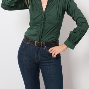 Peut inclure: Une chemise boutonnée vert foncé à manches longues avec un col et des poignets à volants, associée à un jean bleu foncé et une ceinture en cuir noir avec une boucle dorée. La chemise présente un corsage ajusté et des manches longues froncées.