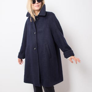 May include: A dark navy wool coat featuring a wide collar and three buttons. The coat has a classic design with a slightly flared shape and long sleeves. The coat is worn with black trousers and boots.