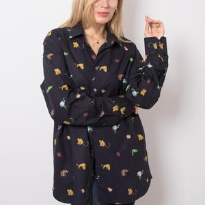 Può includere: Una camicia a maniche lunghe con bottoni in blu navy scuro, caratterizzata da una stampa all-over di piccoli animali colorati, tra cui leoni, elefanti e struzzi. La camicia presenta un colletto classico, un'abbottonatura frontale e polsini con bottoni.