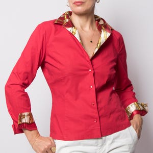 May include: A bright red button-down shirt featuring a contrasting collar and cuffs with a gold and brown chain-link pattern. The shirt has long sleeves, a classic collar, and a buttoned front placket, paired with plain white trousers.