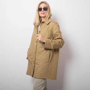 May include: A tan-colored coat with a button-up front, collared neckline, and long sleeves. The coat has two front pockets and is worn over a white top and tan pants. The person is wearing sunglasses.