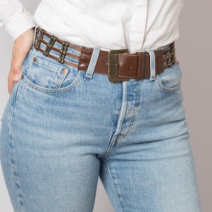 May include: Brown leather belt with a rectangular brass buckle. The belt features a woven design with brass accents. The belt is worn with light blue denim jeans and a white button-down shirt.