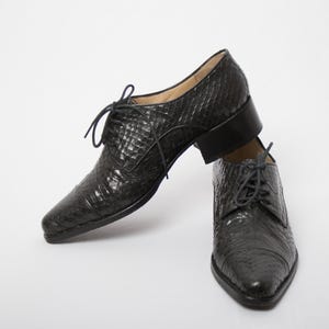 May include: A pair of black leather dress shoes with a textured, reptile-like pattern. The shoes have a pointed toe, lace-up closure, and a small heel. The shoes are set against a white background.