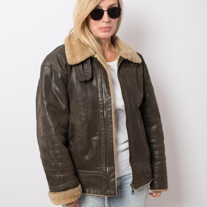 May include: A brown leather jacket with a shearling collar and cuffs. The jacket has a zipper closure and a classic aviator style. The model is wearing sunglasses and light blue jeans.