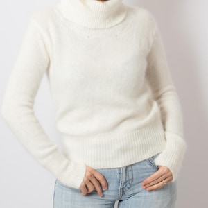 Può includere: Un maglione a collo alto color crema con maniche lunghe. Il maglione ha una trama a coste e una vestibilità leggermente ampia. La modella indossa jeans azzurri. Un capo classico e versatile.