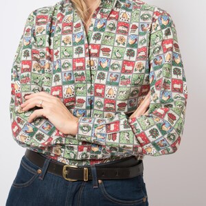 Puede incluir: Una camisa de manga larga con botones y un colorido patrón de cuadrícula con ilustraciones de granja que incluyen gallos, ovejas, graneros y girasoles. La camisa tiene un forro de cuello azul marino y está metida dentro de unos vaqueros azules.