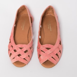 May include: A pair of pink suede flats featuring a woven lattice design across the toe area and an open-toe construction. The interior footbed is tan and displays the gold-stamped text "Les Garçonne" with the year "since 1954" printed clearly.