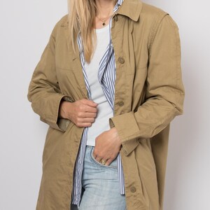 May include: A tan, button-down jacket with a collared neckline. The jacket is worn over a white t-shirt and a blue and white striped shirt. The person is wearing light blue jeans and sunglasses.