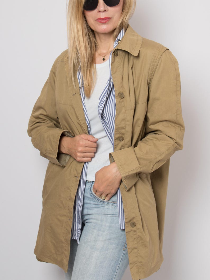 May include: A tan, button-down jacket with a collared neckline. The jacket is worn over a white t-shirt and a blue and white striped shirt. The person is wearing light blue jeans and sunglasses.