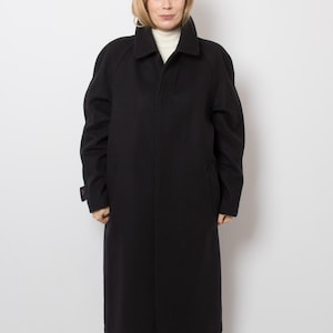 May include: A long, black, heavy-weight coat featuring a structured collar, long sleeves, and a concealed front button placket. The coat has a straight, full-length silhouette with visible side pockets and a clean, solid-colored finish throughout.