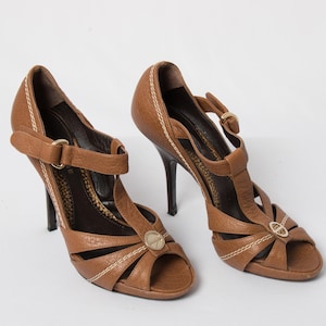 May include: A pair of tan leather high-heeled sandals featuring a T-strap design, an open-toe front, and a gold-toned circular emblem at the center. The shoes include a buckled ankle strap, contrast white stitching along the edges, and a dark interior.