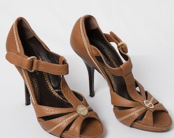 Puede incluir: Un par de sandalias de tacón alto en cuero color canela con diseño en T, punta abierta y un emblema circular dorado en el centro. Los zapatos incluyen una correa de tobillo con hebilla, costuras blancas en contraste y un interior oscuro.