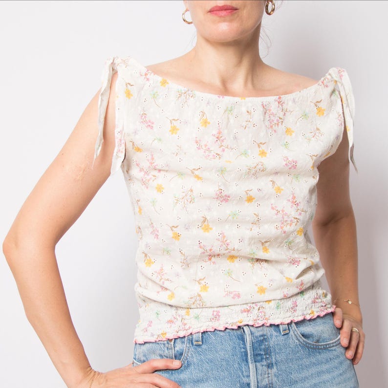May include: A cream-colored eyelet top with a floral pattern in pink and yellow. The top has a square neckline, tie straps, and a pink ruffled hem. The model is wearing blue jeans.