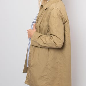 May include: A tan-colored, long-sleeved jacket with a collared neckline. The jacket is buttoned and appears to be made of a lightweight material. A blue and white striped shirt is visible underneath. The jacket is worn over light-wash jeans.