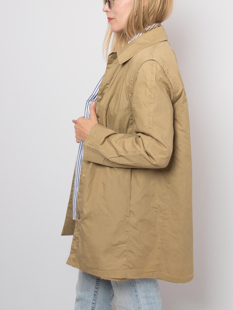 May include: A tan-colored, long-sleeved jacket with a collared neckline. The jacket is buttoned and appears to be made of a lightweight material. A blue and white striped shirt is visible underneath. The jacket is worn over light-wash jeans.
