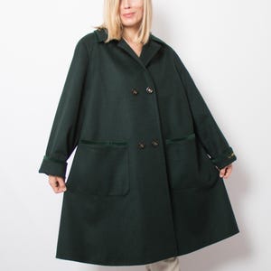 May include: A dark green double-breasted coat with two front pockets and a notched collar. The coat has a slight A-line shape and features dark green velvet trim on the pockets and cuffs. The coat is buttoned up and appears to be made of a wool blend.