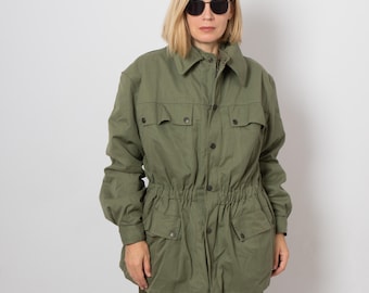 May include: An olive green jacket with a button-up front, four front pockets, and a drawstring waist. The jacket has long sleeves and a collared neckline. The person in the image is wearing sunglasses.