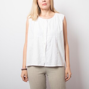 May include: A sleeveless white button-down blouse featuring vertical pintuck pleats along the front placket and a round neckline. The garment is paired with light beige trousers that have a visible woven texture and a straight-leg cut.