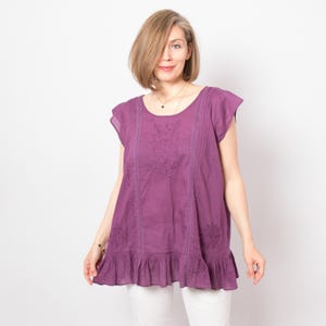 May include: A plum-coloured tunic top with short sleeves and a ruffled hem. The top features embroidered floral designs and vertical stitched panels. The model is wearing the top with white trousers. The top is made of a lightweight fabric.