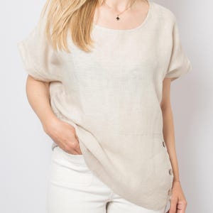 May include: A light beige linen top with short sleeves and a rounded neckline. The top features a buttoned side detail and is paired with white pants. The outfit is simple and elegant, suitable for casual wear.