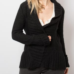 Op de afbeelding: Een zwarte gebreide cardigan met een sjaalkraag. De cardigan heeft een knoopsluiting en een uitlopende zoom. De mouwen zijn lang en de stof lijkt een dikke, getextureerde brei te zijn. De cardigan wordt over een witte top gedragen.