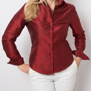 May include: A long-sleeved, button-down shirt in a rich, red color. The shirt has a classic collar and cuffs, and appears to be made of a lustrous fabric. The model is wearing white pants. The shirt is a classic style.