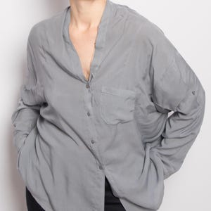 May include: A light gray button-down shirt with a single chest pocket. The long-sleeved shirt has a mandarin collar and is partially unbuttoned. The model is wearing gold hoop earrings and black pants.