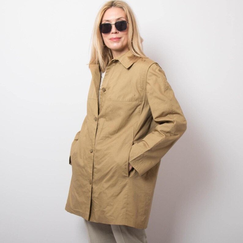 May include: A tan-colored, button-down coat with a collared neckline and pockets. The coat is worn by a person wearing sunglasses. The coat is a neutral color and appears to be made of a lightweight fabric.