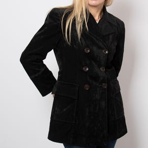 Puede incluir: Un blazer negro de terciopelo con doble botonadura, solapa de muesca, mangas largas con ribete decorativo en los puños y dos grandes bolsillos con solapa en las caderas. La chaqueta se cierra con seis botones oscuros en dos filas verticales.