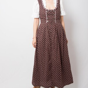 May include: A brown floor-length dirndl dress featuring a small white floral pattern, a square neckline with pink trim, and silver-toned metal bodice hooks. The dress is worn over a white short-sleeved blouse with a ruffled collar and puff sleeves.