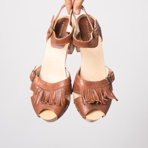 May include: A pair of brown leather clogs with fringe detailing. The open-toe sandals have wooden soles and adjustable ankle straps with buckles. The heels are approximately 3 inches high.