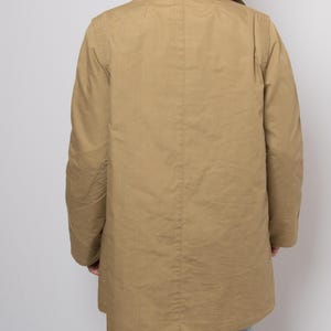 May include: A tan-colored coat with a collared neckline and long sleeves. The coat is buttoned and appears to be made of a lightweight fabric. The person is wearing light blue jeans.