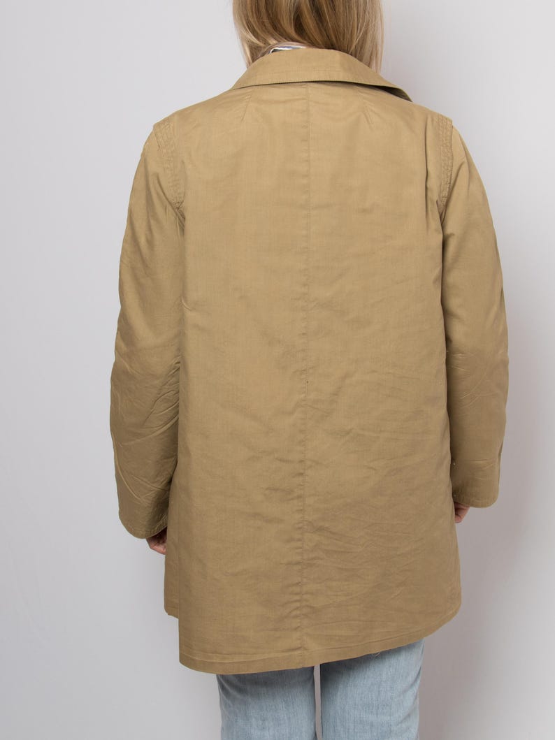 May include: A tan-colored coat with a collared neckline and long sleeves. The coat is buttoned and appears to be made of a lightweight fabric. The person is wearing light blue jeans.