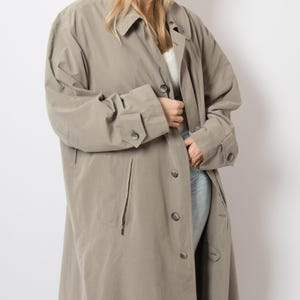 May include: A taupe-coloured trench coat featuring a classic collar, long sleeves with buttoned cuffs, and a front button closure. The coat includes two vertical zippered pockets on the front and a smooth, solid-coloured fabric construction.