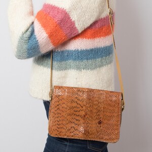 May include: A rectangular crossbody bag featuring a textured brown snakeskin pattern with a flap closure and a thin tan shoulder strap. The bag is worn against a cream-coloured knit sweater with horizontal stripes in shades of orange, pink, and blue.