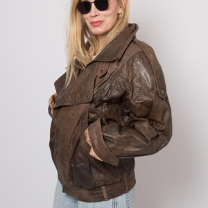 May include: A brown leather jacket with a bomber style design. The jacket has a wide collar, multiple button details, and a ribbed waistband. The jacket is worn with light blue jeans and sunglasses.