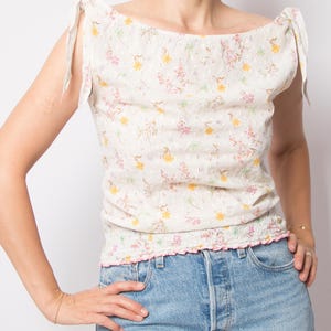 May include: A cream-colored, floral-print top with a scoop neckline and tie details on the shoulders. The top features a delicate pattern of yellow and pink flowers and a ruffled hem. The model is wearing blue jeans.