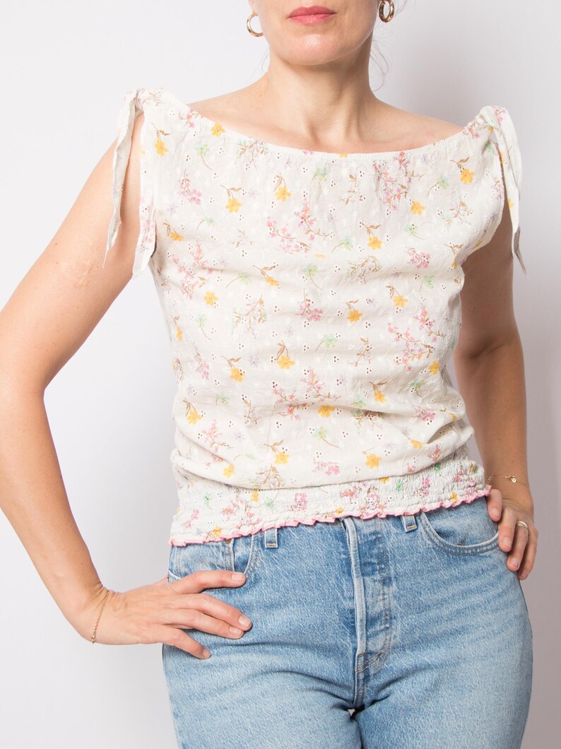 May include: A cream-colored, floral-print top with a scoop neckline and tie details on the shoulders. The top features a delicate pattern of yellow and pink flowers and a ruffled hem. The model is wearing blue jeans.