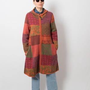 Puede incluir: Un abrigo con capucha y cremallera de manga larga que presenta un diseño de patchwork con varios patrones en tonos naranja, rojo, verde y morado. La prenda tiene una cremallera frontal completa, una capucha y dos bolsillos laterales.