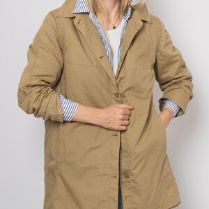 May include: A tan, button-down coat with a collared neckline. The coat is worn over a white shirt and a blue and white striped shirt. The coat has pockets and is knee-length. The person is wearing sunglasses.