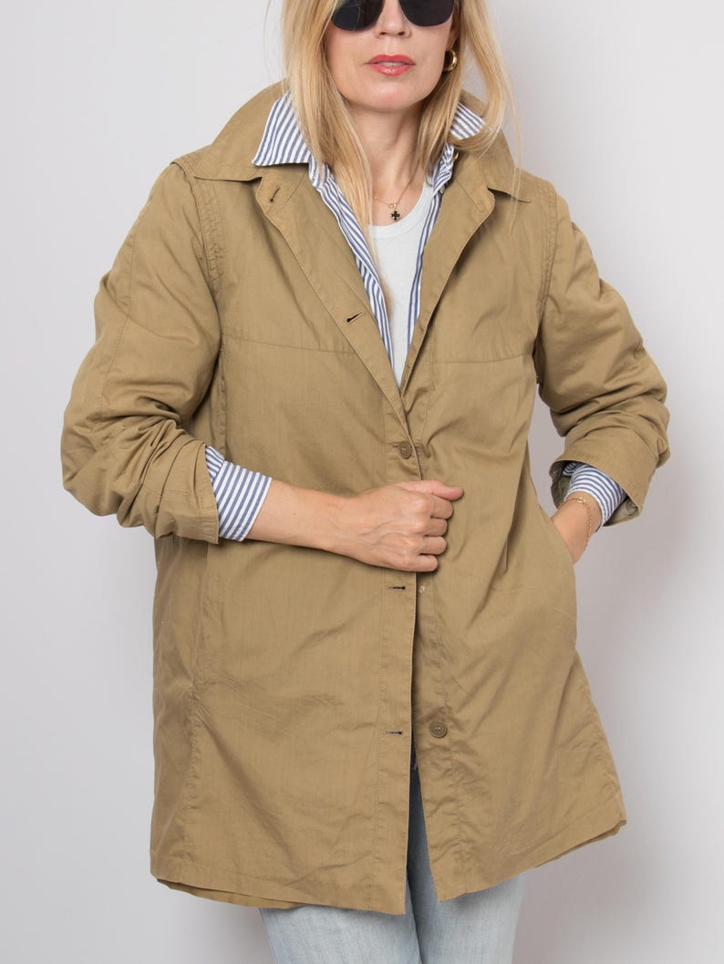 May include: A tan, button-down coat with a collared neckline. The coat is worn over a white shirt and a blue and white striped shirt. The coat has pockets and is knee-length. The person is wearing sunglasses.