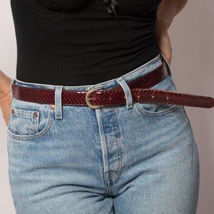 May include: A burgundy snakeskin-patterned belt with a gold-coloured buckle is worn with light blue denim jeans. The belt is the focal point, adding texture and colour to the outfit. The jeans have a classic five-pocket design.