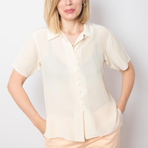 Könnte beinhalten: Ein cremefarbenes kurzärmeliges Hemd mit Knöpfen. Das Hemd hat einen klassischen Kragen und eine Reihe kleiner Knöpfe auf der Vorderseite. Der Stoff wirkt leicht und leicht durchscheinend.