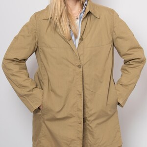 May include: A tan-colored, button-down jacket with a collared neckline and long sleeves. The jacket has two front pockets and is worn over a striped shirt. The model is wearing sunglasses.