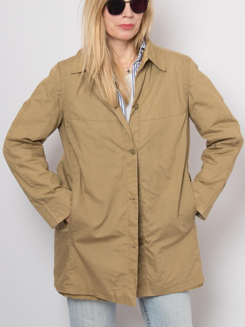 May include: A tan-colored, button-down jacket with a collared neckline and long sleeves. The jacket has two front pockets and is worn over a striped shirt. The model is wearing sunglasses.