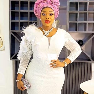 May include: A woman wearing a white lace dress with a ruffled shoulder and a pink head wrap. She is holding a small silver clutch.