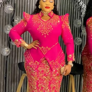May include: A vibrant fuchsia-colored formal dress with intricate gold embroidery. The dress features long sleeves, a fitted bodice, and a floor-length skirt. The design includes shoulder embellishments and a matching gold choker.