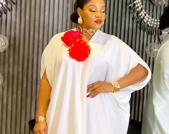 May include: A white, flowing dress with a V-neckline and short sleeves. The dress features a large red floral accent at the neckline and is accessorized with gold jewelry. The model is wearing gold sandals.